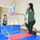 A gymnastics class is taking place in an indoor gym facility. A young toddler in a colorful leotard is standing on a low horizontal bar apparatus while a staff member kneels nearby supervising. A second young child in a black and teal leotard watches from behind, and additional participants are visible in a mirror reflection in the background.