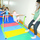 A gymnastics class for young children is taking place in an indoor gymnasium. A small child in a colorful leotard is performing on a low horizontal bar while an adult instructor in green and black attire observes closely. Additional children in leotards are visible in the background practicing near ballet barres.