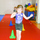 A young girl wearing a purple and black gymnastics leotard is walking barefoot along a low balance beam on a red gymnastics mat. She has her arms outstretched for balance and appears focused on the task. The setting is an indoor gymnastics facility with colorful foam equipment and training cones visible.