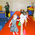 A children's gymnastics or movement class taking place in an indoor gym facility. Three young girls are navigating a balance beam or floor course marked with colorful cones, while an adult instructor in camouflage attire supervises from behind. The space is equipped with foam mats, balance beams, and colorful play equipment.