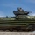 Balikatan 2019: Beach assault with 3/6 The image shows a military amphibious assault vehicle (AAV), likely an AAV-7A1, painted in green and brown camouflage pattern. A single soldier wearing a helmet and goggles is visible in the open commander's hatch at the top of the turret. The vehicle appears to be in motion, photographed against a partly cloudy blue sky.