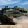 Balikatan 2019: Beach Assault with 3/6 An amphibious assault vehicle (AAV7A1) marked '001' is moving at speed along a dirt trail, kicking up a dust cloud from its tracks. Multiple crew members are visible in open hatches wearing helmets and combat gear. The vehicle is painted in olive drab and brown camouflage and appears to be engaged in a military training exercise.