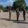 Balikatan 2019: 3rd Battalion, 6th Marine Regiment Promotion A military ceremony is taking place on an outdoor parade ground, likely a change of command or award ceremony involving U.S. Marines in MARPAT camouflage uniforms. Three principal figures stand in the foreground — one holding a guidon, one at attention, and one reading from a document — while formations of troops stand at attention in the background. The setting appears to be an overseas or joint training location based on the mixed troop formations and tropical vegetation.