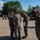 Balikatan 2019: 3rd Battalion, 6th Marines Promotion A military ceremony or formation is taking place at an outdoor base or training area. Several personnel in MARPAT (Marine digital camouflage) uniforms are gathered, with one individual appearing to receive a commendation or pin from a senior officer. A guidon bearer stands to the left holding a red/maroon unit flag.