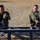 Balikatan 2019: Marines with 3/6 Interact with Philippine Marines Two military personnel in camouflage uniforms are seated on a metal frame structure outdoors, both smiling and laughing. They appear to be taking a break during physical training or a fitness exercise on a dry, open field. The relaxed and jovial demeanor suggests a moment of camaraderie between the two servicemembers.