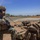 Balikatan 2019: Air Assault with 3rd Battalion, 6th Marines Two U.S. Marines in full combat gear are positioned near what appears to be a runway or airstrip in a tropical environment. The Marine on the left is using a radio handset while armed with a rifle, while the Marine on the right is managing equipment and a large tactical rucksack. Additional personnel are visible in the background in prone positions.