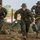 Marines with 3rd Battalion, 6th Marine Regiment Practice their Craft Three military personnel in MARPAT woodland camouflage uniforms are moving rapidly across rocky terrain near a building. They are carrying what appears to be a mortar system and associated equipment including an ammunition case and baseplate. The setting appears to be a training exercise or tactical operation in an arid, semi-rural environment.