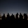 Balikatan 2019: Morning accountability with 3rd Battalion, 6th Marines A group of silhouetted figures stands against a twilight sky transitioning from deep blue to orange near the horizon. At least one figure appears to be carrying a long firearm, visible as a silhouette. The image is a dramatic dusk or dawn scene with approximately 10-12 individuals positioned on elevated ground.