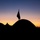 Enjoying the Sunrise during Balikatan 2019 A silhouetted scene captured at dusk or dawn showing multiple dome-shaped tents with a flag mounted on the central tent. The warm orange and yellow horizon contrasts with the deep blue upper sky, creating a dramatic twilight backdrop. A lone human silhouette is faintly visible among the tents in the middle ground.
