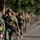 Balikatan 2019: 3/6 room clearing drill, multi-national training with the AFP A large column of military personnel from at least two distinct forces are marching in formation along a paved road lined with large trees. The troops appear to be participating in a joint military exercise or ceremonial march, carrying rifles and wearing different camouflage uniforms. A flag or guidon is visible near the front of the formation.