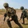 Balikatan 2019: 3/6 Marines conduct Multi-National Training with the AFP Two soldiers in different camouflage uniforms are moving tactically across an open, dry field during what appears to be a military training exercise. The soldier in the foreground wears MARPAT (Marine pattern) camouflage consistent with U.S. Marines, while the soldier in the background wears a different camouflage pattern. Both are armed and moving in a coordinated low-profile advance.