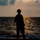 Balikatan 2019: Marines with 3/6 Kilo Company during a sunset in the Philippines A silhouetted figure stands at the edge of a shoreline during sunset or sunrise, facing the open sea. The person is wearing a hat and appears to be holding an object, possibly a fishing rod or walking stick. The warm golden light reflects off the water's surface, creating a dramatic and contemplative atmosphere.