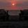 Balikatan 2019: Marines with 3/6 Kilo Company during a sunset in the Philippines Two military personnel in helmets and tactical gear are silhouetted against a sunset over a body of water, standing beside two light tactical utility vehicles on a sandy beach. The scene is dramatically backlit by a red-orange sun low on the horizon. The image has a strong compositional symmetry with one soldier and one vehicle on each side.
