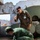 Humanitarian Mine Action increases demining capacity in Morocco Two military personnel are engaged in what appears to be a training exercise at a deteriorated facility. One individual in US Marine Corps MARPAT camouflage uniform with a name badge observes while a second person in solid olive/green uniform kneels on the ground handling equipment near a pole. The setting includes damaged concrete structures, razor wire, and what appears to be industrial or military equipment in the background.