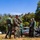 Humanitarian Mine Action increases demining capacity in Morocco A group of military personnel, including one individual in US Marine Corps MARPAT camouflage and several others in solid olive/green uniforms, are gathered outdoors examining what appears to be an unexploded ordnance or rocket on the ground. The scene appears to be a training or EOD (Explosive Ordnance Disposal) exercise in a field environment. Abandoned vehicles and cylindrical objects are visible in the background.