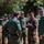 Humanitarian Mine Action increases demining capacity in Morocco A group of military personnel in an outdoor setting appear to be engaged in a briefing or discussion. The group includes individuals in green military uniforms and one person in US Marine Corps digital camouflage (MARPAT) pattern. The setting appears to be a training area or military compound with trees and a white wall visible in the background.
