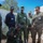 Humanitarian Mine Action increases demining capacity in Morocco A group of military personnel from multiple nations are gathered outdoors observing a demonstration involving a rocket or projectile on the ground. One individual in dark green uniform and a cap marked 'G.S.I.' is crouching and gesturing toward the munition, appearing to explain or demonstrate its components. A U.S. Marine in MARPAT camouflage and personnel in various other military uniforms, including a navy blue uniform and olive green uniforms, observe attentively.