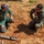 Humanitarian Mine Action increases demining capacity in Morocco Two military personnel are crouching on sandy ground examining what appears to be inert training ordnance or demonstration munitions. One individual wears green coveralls with a cap bearing Arabic script and a badge, while the other wears US Marine Corps digital camouflage (MARPAT) uniform. The setting appears to be an outdoor training or demonstration area, suggesting a joint military exercise or EOD (Explosive Ordnance Disposal) training event.