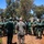 Humanitarian Mine Action increases demining capacity in Morocco A military briefing or training session is taking place outdoors, with one individual in US Marine Corps digital camouflage uniform addressing a large group of personnel wearing olive/dark green uniforms. The setting appears to be a training facility or base with red dirt ground, eucalyptus trees, and simple structures in the background. The gathering suggests a multinational military training exercise or advisory mission.