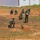 Humanitarian Mine Action increases demining capacity in Morocco A group of personnel in green military/EOD uniforms are conducting what appears to be an explosive ordnance disposal (EOD) or demining training exercise in an open dirt field. One individual is kneeling close to a partially buried cylindrical object resembling an unexploded ordnance (UXO) or shell casing, while others observe from a safe distance. A supervisor or instructor appears to be holding documents and overseeing the exercise.