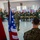 Humanitarian Mine Action increases demining capacity in Morocco A U.S. Marine in MARPAT camouflage uniform is addressing a large group of soldiers wearing olive green uniforms in a classroom setting. Two flags are visible in the foreground — the American flag and what appears to be the Moroccan flag — suggesting a joint military training or exchange event. The audience appears engaged and attentive, with some smiling.