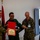 Humanitarian Mine Action increases demining capacity in Morocco A military ceremony is taking place in an indoor setting where a certificate or letter of commendation is being presented. Four military personnel are present, including what appear to be U.S. Army and U.S. Marine Corps members alongside a foreign military officer in green uniform. The atmosphere is celebratory, with participants smiling and laughing.