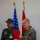 Humanitarian Mine Action increases demining capacity in Morocco Two military personnel are posing together during what appears to be a formal recognition ceremony. One individual in green military uniform is receiving a certificate or letter of commendation in a red folder from another individual wearing US Marine Corps MARPAT camouflage. Two flags — the United States flag and what appears to be the Jordanian flag — are visible in the background.