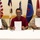 Three individuals are seated at a white-draped table holding up signed documents in what appears to be an official signing ceremony. The group includes a U.S. Army soldier in camouflage uniform on the left, a civilian in a red shirt in the center, and an individual wearing a traditional lei necklace on the right. Multiple flags are displayed in the background, suggesting a formal governmental or military agreement.