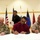 A formal signing ceremony is taking place at a table with three seated individuals and one standing person in the background. A U.S. Army soldier in camouflage uniform is seated on the left alongside two civilians, all appearing to sign documents. The setting appears to be a military facility, indicated by unit insignia on the walls including a 'Pride of the Pacific' patch and a Roman numeral IX emblem.