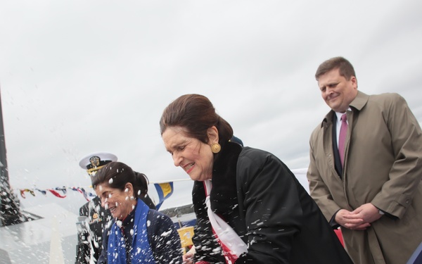 A naval ship christening ceremony is taking place, with a woman in a black coat smashing a bottle against the bow of a warship, causing champagne or liquid to spray dramatically. Several dignitaries and officials are present on the deck, with a naval officer in white uniform visible in the background. The ship appears to be a modern U.S. Navy destroyer or similar warship, decorated with ceremonial bunting in red, white, and blue.