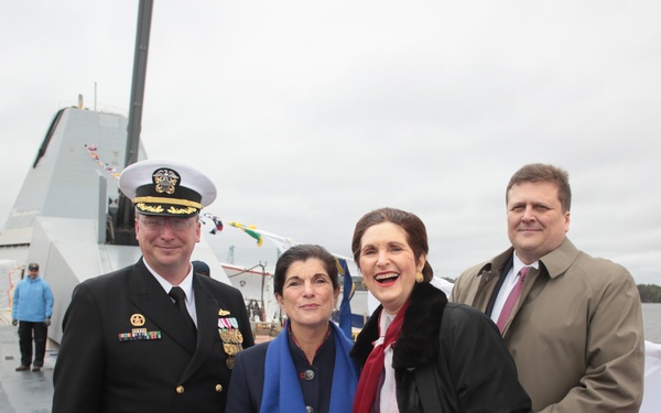 Four individuals pose for a photograph on the deck of a naval vessel during what appears to be a commissioning or christening ceremony. A senior U.S. Navy officer in dress uniform stands alongside three civilians, with colorful bunting and a large warship's superstructure visible in the background. The overcast weather and formal attire suggest an official military ceremony.
