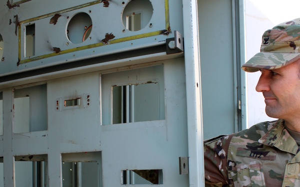 A U.S. Army Colonel in OCP (Operational Camouflage Pattern) uniform is examining a large, weathered military equipment panel or control console. The panel appears to be a decommissioned or legacy military electronic/electrical equipment housing with multiple circular cutouts, rectangular openings, and numerous small holes. The soldier is looking intently at the panel, possibly conducting an inspection or assessment.