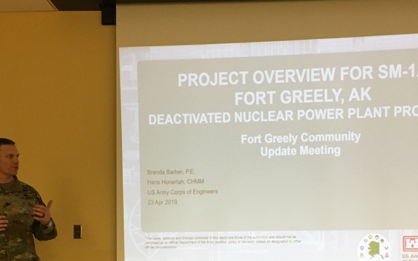 A U.S. Army soldier in camouflage uniform is presenting at a community update meeting about the SM-1A Fort Greely, Alaska Deactivated Nuclear Power Plant Program. The presentation slide, dated April 23, 2019, is projected on a large screen and credits Brenda Barber, P.E. and Hans Honerlah, CHMM from the U.S. Army Corps of Engineers. The setting appears to be an indoor meeting room at a military installation.