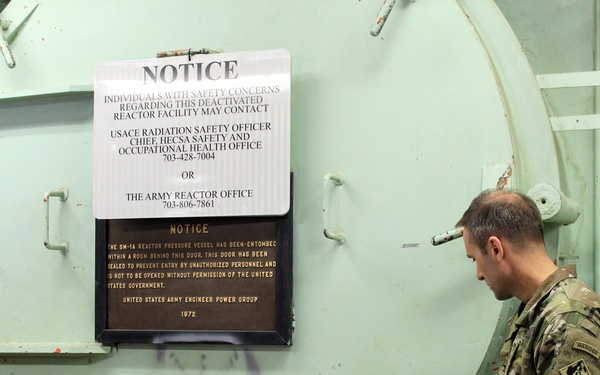 A U.S. Army soldier in camouflage uniform examines a large green industrial door or hatch at a deactivated nuclear reactor facility. Two notices are posted on the structure: one directing individuals with safety concerns to contact the USACE Radiation Safety Officer and the Army Reactor Office, and a second plaque from 1972 indicating the SM-1A reactor pressure vessel has been entombed behind the door. The facility appears to be a decommissioned U.S. Army nuclear reactor site.