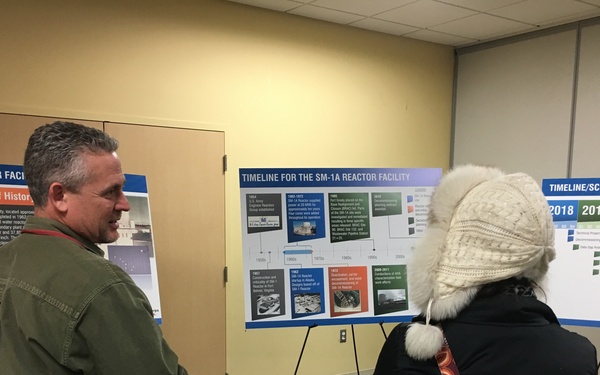 Two individuals are viewing informational display boards at what appears to be a public meeting or open house about the SM-1A Reactor Facility. The boards include a brief history panel, a timeline for the SM-1A Reactor Facility, and a Timeline/Schedule board showing years 2018-2021. The setting is an indoor conference or meeting room with carpeted flooring.