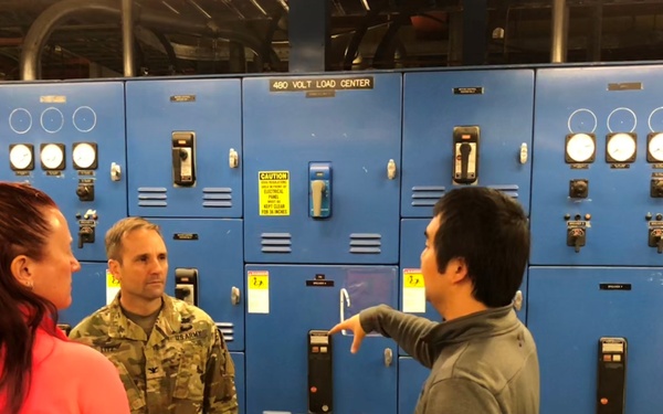 Three individuals are standing in front of a large blue 480-volt electrical load center panel. One person is wearing a U.S. Army camouflage uniform, while the others are in civilian clothing. The person on the right appears to be pointing at or explaining something on the electrical panel to the others.