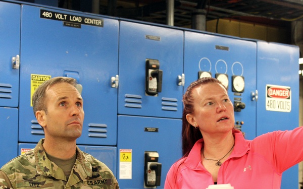 A U.S. Army Colonel in camouflage uniform stands beside a woman in a pink top who is pointing toward something off-frame, apparently giving a tour or briefing. They are standing in front of large blue electrical control panels labeled '480 Volt Load Center' with a 'Danger 480 Volts' warning sign visible. The setting appears to be an industrial or military facility electrical room.