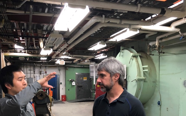 Two men are conversing inside an industrial facility, likely a manufacturing plant or mechanical room. One man is gesturing with his hand while explaining something, while the other holds what appears to be technical drawings or blueprints. The space contains large industrial equipment including what appears to be a pressure vessel or autoclave.