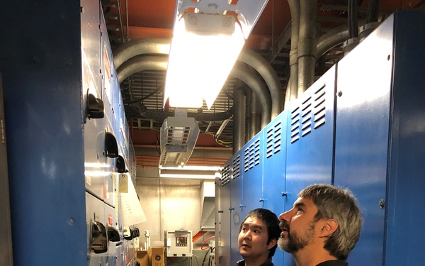 Two men are standing in a narrow industrial electrical room or switchgear corridor, both looking upward, apparently inspecting overhead infrastructure. The space is flanked by large blue electrical panels or switchgear cabinets on both sides. One man holds papers and the other holds what appears to be a pen or stylus, suggesting a formal inspection or assessment is underway.