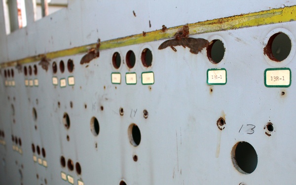 The image shows an abandoned or decommissioned industrial control panel or electrical switchboard in a state of significant deterioration. The metal panel surface is heavily rusted and stained, with numerous circular cutouts where instruments or switches have been removed. Green-bordered labels and handwritten numbers remain visible, suggesting this was once an organized, numbered control system.
