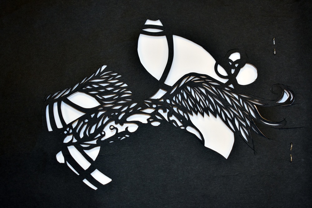 The image depicts an intricate black paper-cut artwork laid on a dark background, featuring a bird — likely a crane or phoenix — in flight with detailed feather work. White paper or backing is visible beneath the cutout, creating strong contrast. Small metal pins or clips are visible to the right side, suggesting the piece is in progress or being assembled.