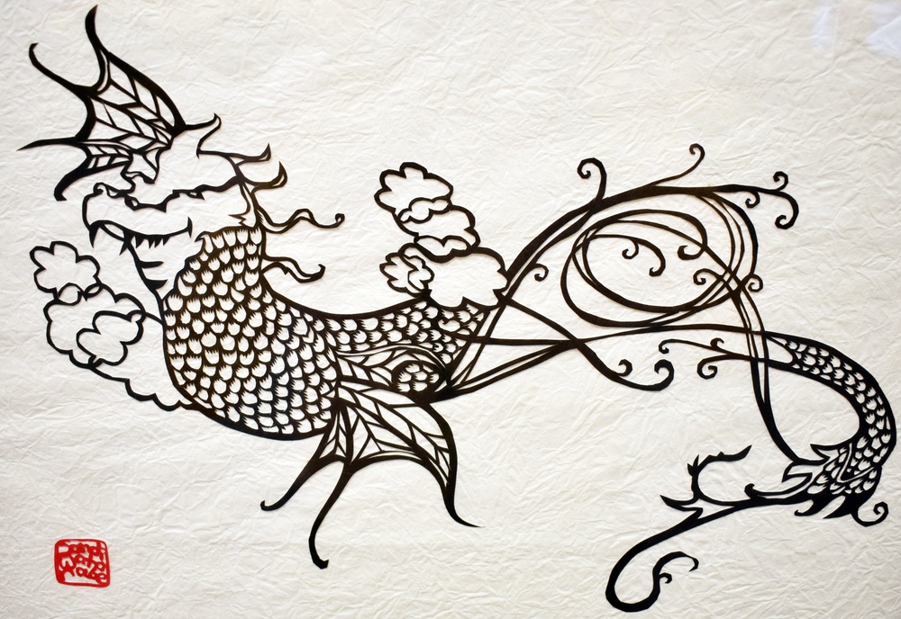 A detailed black ink artwork on textured white paper depicting a stylized phoenix or mythical bird in flight, rendered in what appears to be a paper-cutting (kirie) or ink illustration technique. The creature features intricate scale patterns, flowing tail feathers rendered as elegant curling lines, and is surrounded by stylized clouds. A small red seal stamp is visible in the lower left corner, suggesting East Asian artistic tradition.