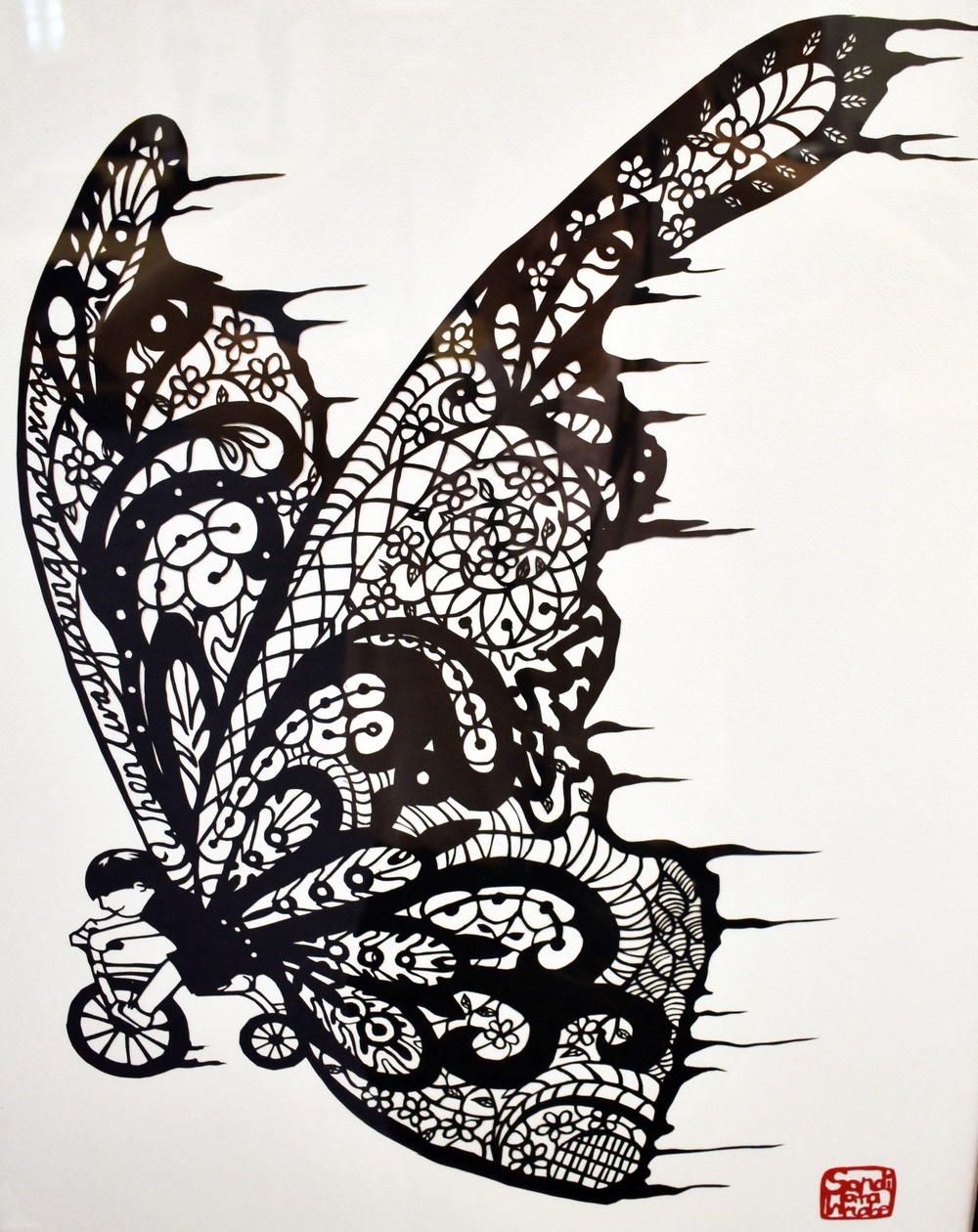 A highly detailed black-and-white paper-cut or ink artwork depicting a large, ornate butterfly wing composed of intricate lace-like patterns including florals, swirls, and geometric designs. At the lower left, a small child figure rides a bicycle, appearing to be propelled or carried by the massive decorative wing. The artwork bears a red artist's seal/stamp in the bottom right corner.