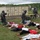 This image depicts a HAZMAT or CBRN (Chemical, Biological, Radiological, Nuclear) emergency response training exercise. Personnel in full protective suits and gas masks are triaging simulated casualties (role players or mannequins) lying on the ground. The scene includes shipping containers, a wheeled stretcher, and an industrial background consistent with a training facility.