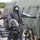 A person wearing full CBRN (Chemical, Biological, Radiological, and Nuclear) protective gear stands with arms outstretched in front of a large military-style green tent structure. The setting appears to be a military or emergency response training exercise, with additional personnel in similar protective suits visible in the background. Various decontamination equipment and infrastructure are visible around the scene.