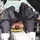 This image depicts a military or emergency response training exercise involving CBRN (Chemical, Biological, Radiological, Nuclear) decontamination procedures. Personnel in full protective suits and respirators are treating a simulated casualty on a stretcher with simulated wounds (moulage makeup). The scene includes decontamination equipment such as buckets and brushes, consistent with a mass casualty drill.