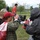 The image depicts what appears to be an emergency response or disaster drill exercise taking place on a grassy outdoor area. Several participants are visible, including individuals wearing gas masks and protective gear, while others appear to be civilian role-players with tagged mesh bags. A person lying on the ground suggests a simulated casualty scenario.