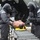 A military or emergency response training exercise depicting a CBRN (Chemical, Biological, Radiological, Nuclear) decontamination scenario. Two personnel in full protective gear including gas masks and gray hazmat suits are processing a simulated casualty (moulage mannequin or actor with simulated wounds) on a roller conveyor system. The setting appears to be an outdoor field decontamination station with tent structures.
