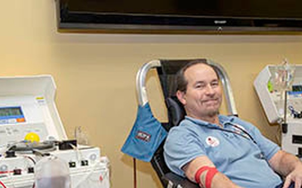 A middle-aged man is seated in a medical recliner chair, smiling, while undergoing an apheresis or blood donation procedure. He is connected to medical equipment via tubing attached to his arm, with a blood pressure cuff visible on his upper arm. The setting appears to be a blood or plasma donation center with multiple apheresis machines visible.
