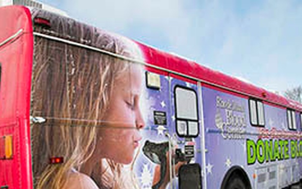 A mobile blood donation bus is parked in a lot, wrapped in colorful vinyl graphics featuring a young girl and the message 'Donate Blood.' The bus belongs to a Blood Center organization and is set up as a mobile donation unit. Safety cones and a sandwich board sign reading 'Donate Blood Today' are placed outside the vehicle.